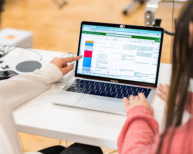 Two people (mostly off-screen) looking at a laptop displaying a spreadsheet full of data. 